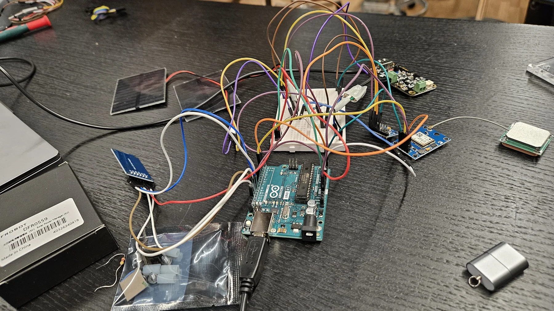 Close view of an Arduino Uno, breadboard wiring, connected modules, and a small solar panel on a workbench.