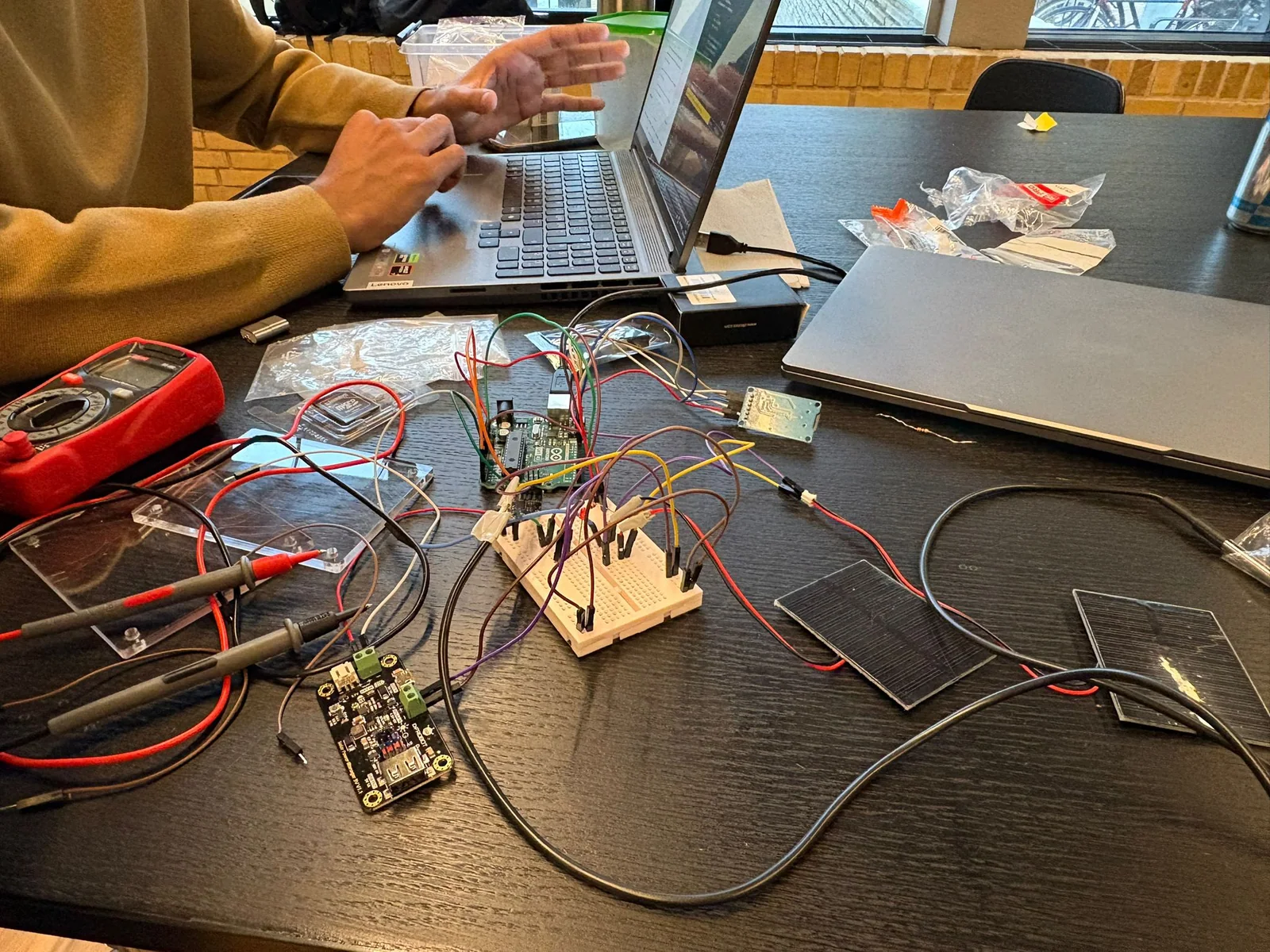 Bench setup with an Arduino board, breadboard wiring, solar panels, multimeter, and laptop.